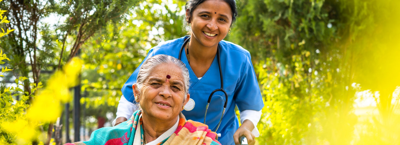 happy smiling Nurse taking senior woman on walk while on wheelchair at hospital garden by looking at camera - concept of caretaker, disability and healthcare