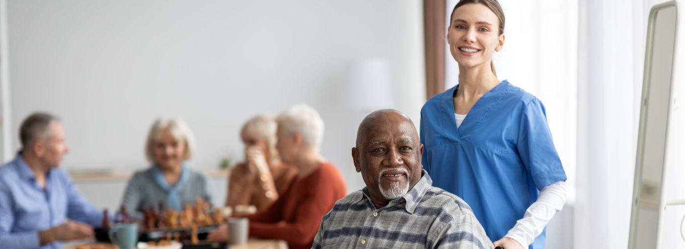 Happy older patient on wheelchair with female nurse smiling at camera, group of senior people sitting on couch on background, nursing home interior, healthcare for elderly people concept